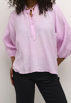Femme portant un chemisier texturé violet clair avec trois boutons et un lien au col et un pantalon noir, les mains sur les hanches.