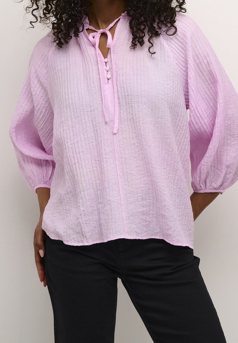 Femme portant un chemisier texturé violet clair avec trois boutons et un lien au col et un pantalon noir, les mains sur les hanches.