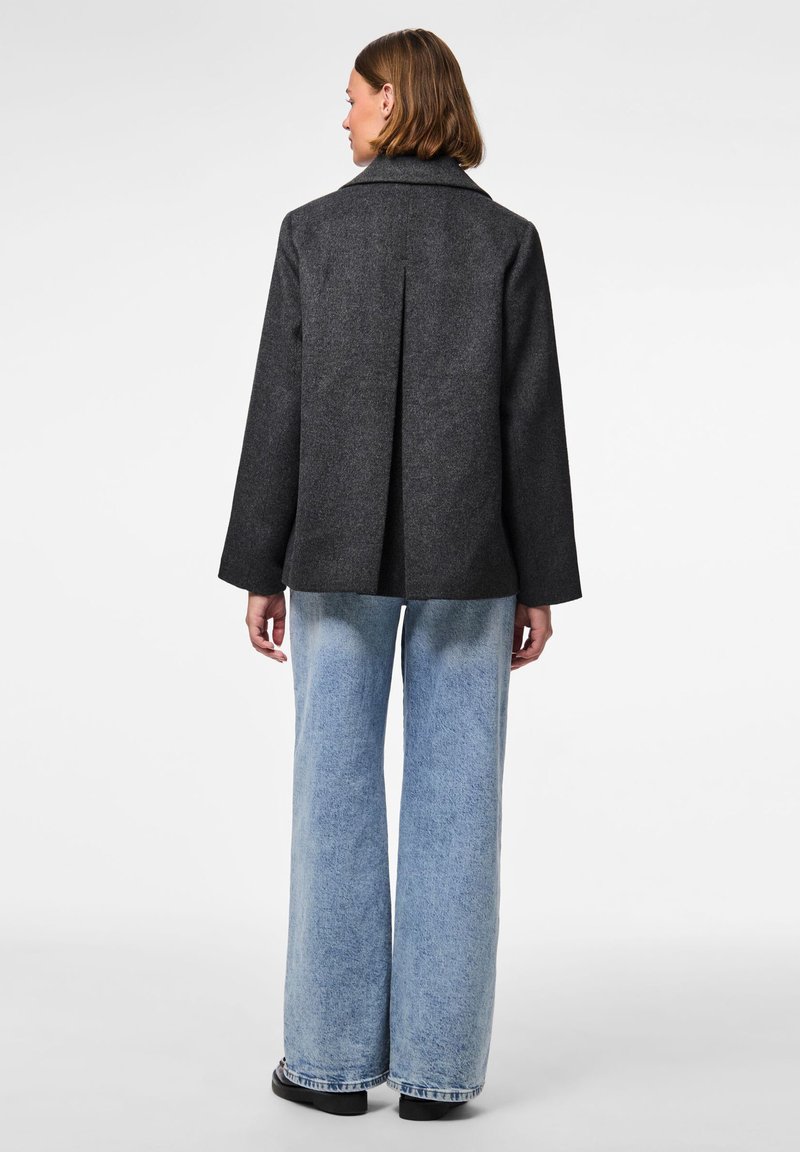 Grå ullkappa med slagkrage och vent bak, tillsammans med vida ljusblå jeans i denim. Svarta platta skor fulländar looken.