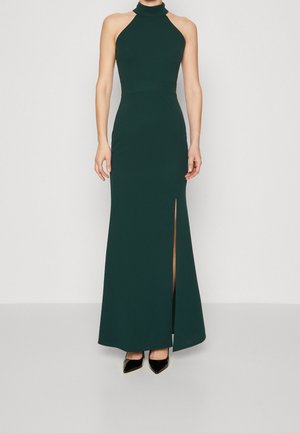 Kvinde iført en mørkegrøn ærmeløs halterneck-kjole med høj slids og sorte spidse hæle, stående foran en ensfarvet baggrund.