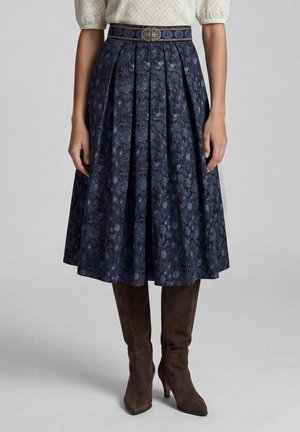 Femme portant une jupe plissée bleu marine à motif floral, longueur genou, avec une ceinture décorative et des bottes en daim marron à talons hauts.