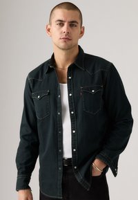 Dark green denim shirt with two chest pockets, silver snap buttons, contrast stitching, and a classic collar, worn over a white tank top.