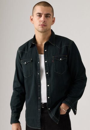 Dark green denim shirt with two chest pockets, silver snap buttons, contrast stitching, and a classic collar, worn over a white tank top.