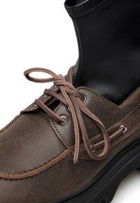 Stivale alla caviglia in pelle marrone con la parte superiore nera liscia. Presenta lacci marroni, occhielli in argento e una suola nera spessa per un supporto aggiunto.