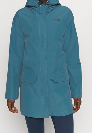 Türkisfarbener knielanger Regenmantel mit Kapuze, Knopf- und Reißverschluss sowie Klapptaschen, getragen über dunklen Leggings.