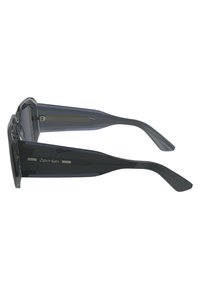 Occhiali da sole Calvin Klein con montatura grigia trasparente, lenti scure e design elegante e angolare. Presentano linee di accento argentate sulle tempie.