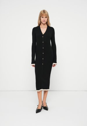Vestido de punto negro con mangas largas, escote en V, botones dorados y un diseño texturizado. Presenta un borde color crema en los puños y el dobladillo.