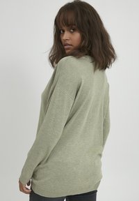 Maglione verde chiaro in un morbido tessuto a maglia, con una vestibilità rilassata, spalle scese e polsini a coste, indossato con jeans scuri.