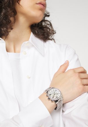 Personne portant une chemise blanche avec une montre-bracelet argentée avec un grand cadran rond et des chiffres romains sur le cadran.