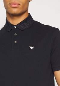 Svart pikétröja i bomull med texturerad krage, två-knapps knäppning och en liten vit logotyp på bröstet.