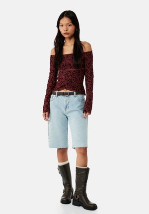 Młoda kobieta w bordowej bluzce z odkrytymi ramionami w kwiatowy wzór, jasnoniebieskich dżinsowych szortach do kolan, ciemnobrązowych botkach i białych skarpetkach, stoi na białym tle.