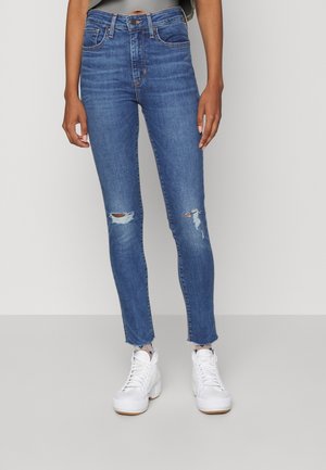 Personne portant un jean skinny bleu déchiré avec des ourlets effilochés et des baskets blanches à plateforme, debout devant un fond uni.