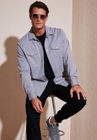 Cămașă gri cu nasturi, având două buzunare la piept, purtată peste o cămașă neagră, combinată cu pantaloni negri și adidași gri, așezat pe un taburet.