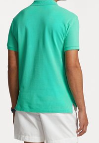 Mies, joka on pukeutunut turquoise-väriseen lyhythihaiseen polo-paitaan ja valkoisiin shortseihin, näkyy takaa ja hänellä on kello vasemmassa ranteessaan.