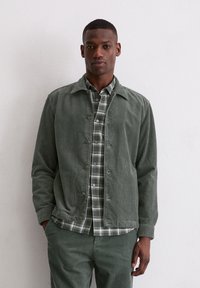 Zielona kurtka ze sztruksu z kołnierzem, zestawiona z zielono-białą koszulą w kratę. Charakterystyczna prążkowana struktura i spodnie w tym samym kolorze.