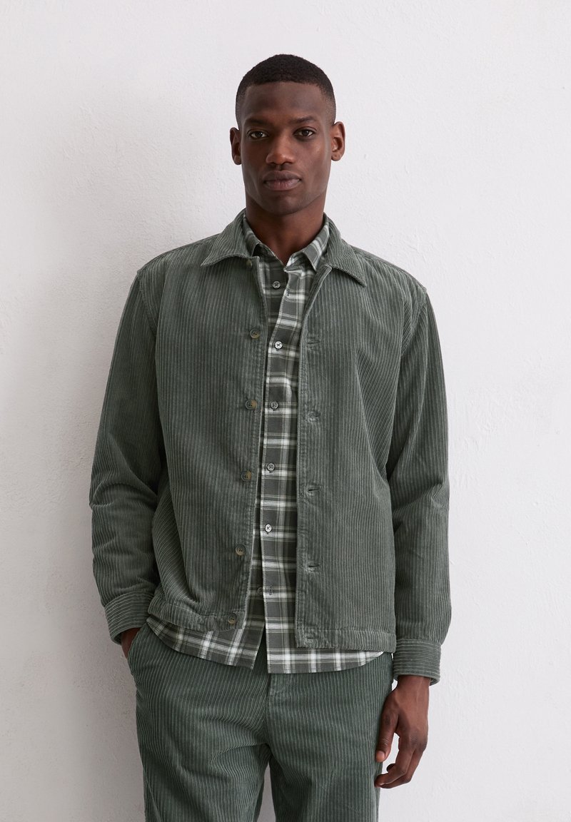 Zielona kurtka ze sztruksu z kołnierzem, zestawiona z zielono-białą koszulą w kratę. Charakterystyczna prążkowana struktura i spodnie w tym samym kolorze.