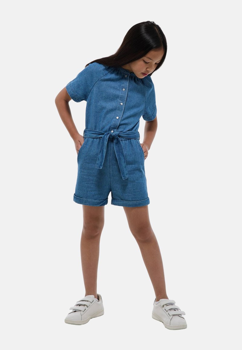 Salopette en denim à manches courtes, col, boutons sur le devant et ceinture à nouer. Comprend des poignets retroussés et une coupe décontractée. Des baskets blanches complètent le look.