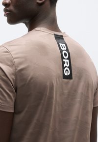 T-shirt camouflage marron avec un col rond et des manches courtes. Présente une étiquette verticale noire avec le texte "BORG" dans le dos.