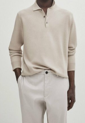 Mann trägt ein beiges Langarm-Poloshirt, das in hellbeige Hosen gesteckt ist, mit Händen in den Taschen.