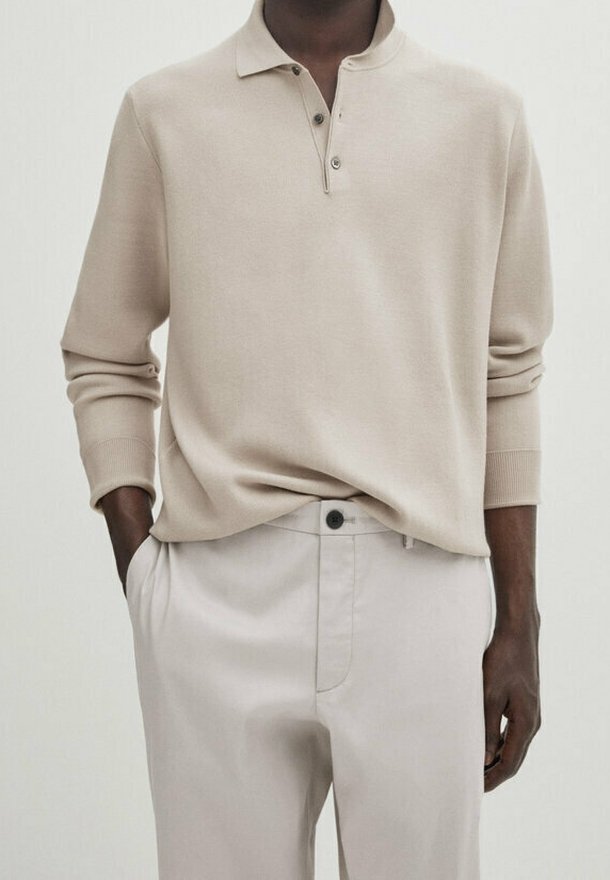 Homme portant un polo à manches longues beige, rentré dans un pantalon beige clair, les mains dans les poches.