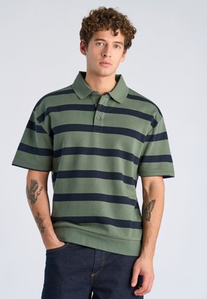 Giovane uomo con capelli ricci che indossa una polo a righe verdi e nere e jeans scuri, in piedi con una mano in tasca contro uno sfondo semplice.