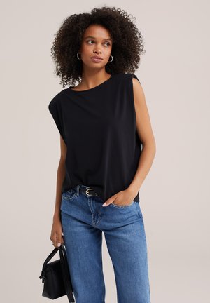 Frau mit lockigem Haar, die ein ärmelloses schwarzes Oberteil und blaue Jeans trägt, hält eine schwarze Handtasche und steht vor einem einfarbigen Hintergrund.