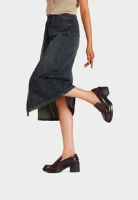 Gonna di denim blu scuro abbinata a scarpe con piattaforma bordeaux lucido, con punta rotonda e sottili accenti verticali.