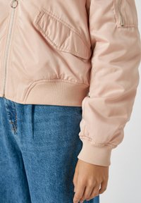 Personne portant une veste bomber rose clair avec une poche zippée et un jean en denim bleu, main détendue le long du corps.