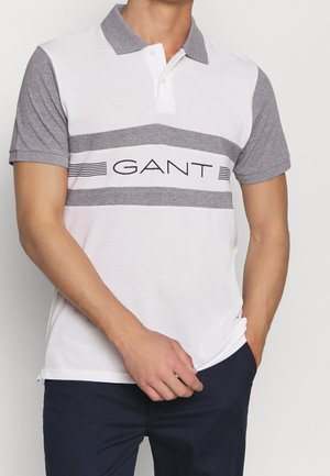 Homme portant un polo à manches courtes blanc et gris avec le logo "GANT" sur la poitrine, associé à un pantalon foncé, une main dans la poche.