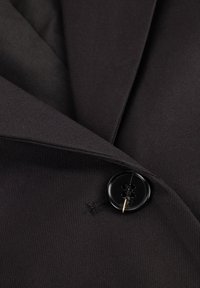 Zwarte stoffen blazer met een ingesneden revers, voorzien van een grote zwarte knoop met vier gaatjes en een subtiele geribbelde textuur.