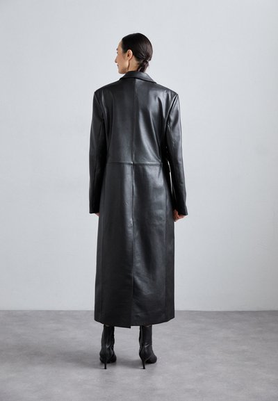 Schwarzer Leder-Mantel mit einer taillierten Silhouette, abgesetztem Kragen und einer Mittelnähten-Detail. Gestylt mit knielangen schwarzen Stöckelschuhen.