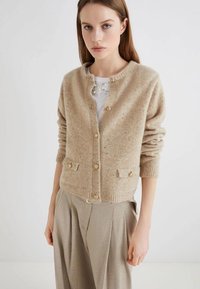 Cardigan in lana beige con collo rotondo, cinque bottoni dorati e due tasche frontali. La trama è morbida con un leggero motivo a macchie.