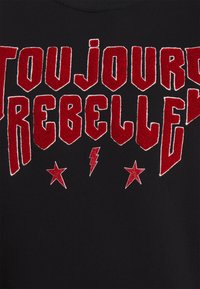Tela negra con texto bordado en rojo intenso que dice "TOUJOURS REBELLE", con estrellas y rayos de brillo como adornos.