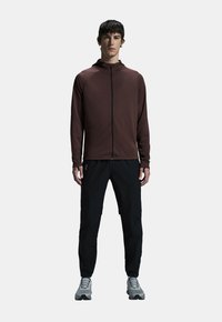 Giacca marrone da uomo con zip e cappuccio, abbinata a pantaloni neri e sneakers grigie. La giacca presenta un tessuto liscio e una branding minima.