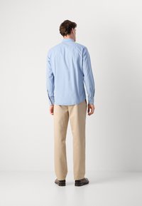 Camisa azul-claro de manga longa com punhos abotoados e pala nas costas, combinada com calças bege e sapatos escuros, exibida de costas.