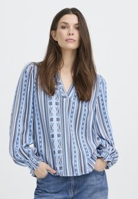 Blouse à rayures bleues et blanches avec col en V, présentant des motifs géométriques et des poignets élastiqués, confectionnée dans un tissu léger.