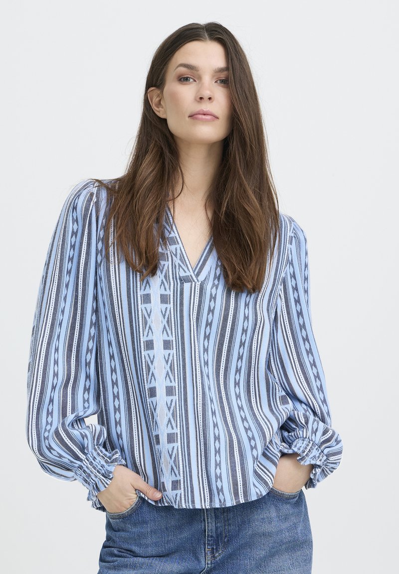 Blouse à rayures bleues et blanches avec col en V, présentant des motifs géométriques et des poignets élastiqués, confectionnée dans un tissu léger.