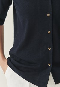 Cămașă bleumarin, cu guler, din material ușor, cu mâneci scurte, având nasturi din lemn și un croi lejer. Materialul pare moale, cu o textură netedă.