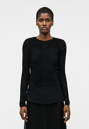 Vrouw die een zwarte top met lange mouwen en geplooide zoom draagt, staand tegen een effen lichte achtergrond, met een neutrale uitdrukking en recht vooruit kijkend.