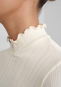 Top color crema con un colletto alto a coste e un bordo ondulato. Il tessuto presenta strisce verticali che conferiscono un aspetto testurizzato.