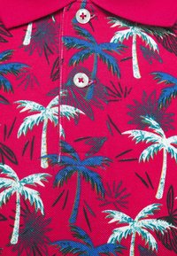 Tissu de polo rouge avec des boutons blancs décorés de motifs de palmiers bleus et blancs et des accents de feuilles noires.