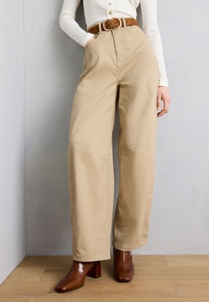 Άτομο που φοράει μπεζ παντελόνι με φαρδιά μπατζάκια και καφέ ζώνη, λευκό μακρυμάνικο τοπ και καφέ μποτάκια με τετράγωνη μύτη και τακούνι, στέκεται σε ξύλινο πάτωμα.