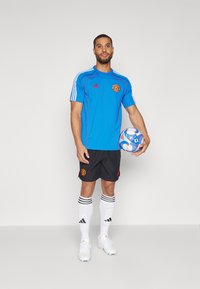 Mėlynas Adidas futbolo marškinėliai su raudonais akcentais, juodi šortai, balti kojinės su juodomis juostelėmis ir baltos sportbačiai, laikantys raštuotą futbolo kamuolį.
