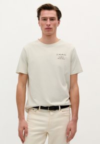 Lichtbeige Baumwoll-T-Shirt mit Rundhalsausschnitt, versehen mit schwarzem Drucktext "SCALPERS SUMMER ESSENTIALS" auf der linken Brust. Getragen mit beigen Hosen.