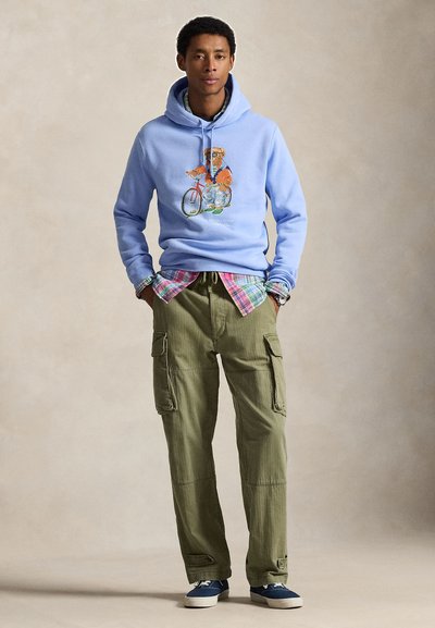 Ανοιχτό μπλε φούτερ με κουκούλα και γραφικό αρκούδας, φορεμένο πάνω από καρό πουκάμισο. Φόρμες cargo ελιάς με πολλές τσέπες. Μπλε αθλητικά παπούτσια.