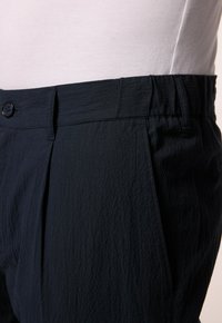 Marinblå byxor med texturerad tyg, plisserad front och knappstängning vid midjan. Detalj med elastisk midja synlig.