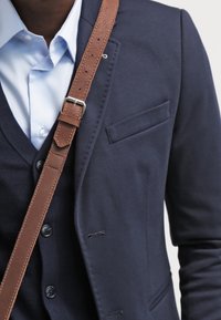 Marinblå blazer med smal passform, med slagkrage, bröstficka och ett brunt läderaxelband över en ljusblå skjorta.