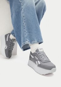 Osoba v svetlomodrých džínsach a bielych ponožkách, ukazujúca šedé a biele platformové tenisky Reebok na jednoduchom pozadí.