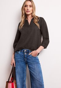 Femme portant un chemisier noir à manches longues et un jean taille haute bleu, tenant un sac fourre-tout multicolore avec des anses marron.