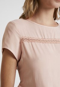 Jasnoróżowa bluzka z krótkim rękawem, z okrągłym dekoltem i ozdobną haftowaną aplikacją na górnej części przodu, wykonana z miękkiego, lekkiego materiału.